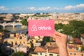 Rome, Italy - May 13, 2018: Person holding Airbnb logo in hand with city in background.