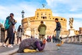 Beggar woman asking alms on her knees on Ponte Sant `Angelo, Bridge of Hadrian full of walking people with Castel Sant `Angelo,