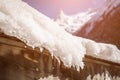 Roof of the house is covered with a thick layer of snow and icicles on the background of snow-capped mountains and bright sun