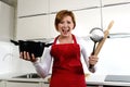 Rookie home cook woman in red apron at home kitchen holding cooking pan and rolling pin screaming desperate in stress