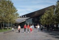 Family group of people walking through pedestrian area in summer sunshine. Modern building and trees in background