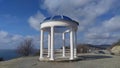 A rotunda with concrete columns stands on top of a hill. There are bushes and trees on the slope. On one side is the sea, on the o