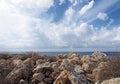 Rough rocks and boulders with dry summer vegetation in front of a calm sea with white sunlit clouds filling the sky