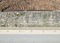 Rough wall made of concrete and stones with a tiled rooftop behind it. Sidewalk and asphalt road in front.