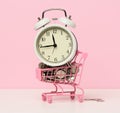 Round alarm clock in a miniature shopping cart with change on a white table. Concept time is money, waste of money and poverty Royalty Free Stock Photo