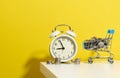 Round alarm clock, miniature shopping cart with coins on a white table. Concept time is money, waste of money and poverty Royalty Free Stock Photo