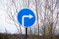 Round blue road sign with white arrow turn right against the backdrop of bare tree branches and sky Royalty Free Stock Photo