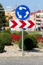 Round blue and white roundabout road sign above red and white striped arrow dangerous turn road sign surrounded with gravel and