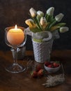 A round candle is lit in a glass candle holder near a vase of white flowers on a wooden table with some strawberries and dry plant Royalty Free Stock Photo