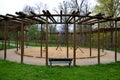 Round pergola, trellises made of wooden poles around the sandpit with play elements for children. around is a path and lawns. benc