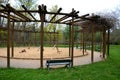 Round pergola, trellises made of wooden poles around the sandpit with play elements for children. around is a path and lawns. benc