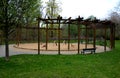Round pergola, trellises made of wooden poles around the sandpit with play elements for children. around is a path and lawns. benc