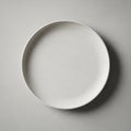 A round, plain white ceramic plate with a smooth texture rests on a similarly textured light gray Royalty Free Stock Photo