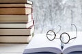 Round reading glasses lie on an open book with white pages next to a stack of multi-colored books on a windowsill