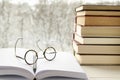Round reading glasses lie on an open book with white pages next to a stack of multi-colored books on a windowsill