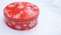 Round, red cookie and baked goods aluminum tin container decorated with white snowflake print pattern, resting on natural snow. Royalty Free Stock Photo