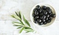 Bowl of Shiny Black Olives on a Rustic White Wooden Surface Next to Olive Branches Royalty Free Stock Photo