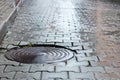 Round steel sewer manhole on wet cobblestone road Royalty Free Stock Photo
