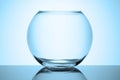 A round transparent empty aquarium stands on a reflective glass surface on a white background. Cold toned blue