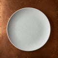 A round, white ceramic plate with subtle line designs sits on a textured copper surface Royalty Free Stock Photo