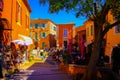 View beyond tree on street with bright yellow, ochre and red French mediterranean houses against blue sky