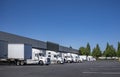 Row of the big rig semi trucks with semi trailers standing at warehouse dock loading cargo for the next freight Royalty Free Stock Photo