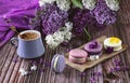 A row of colorful macaroon desserts on the table and a gray cup of coffee. Purple bouquet of lilac flowers Royalty Free Stock Photo