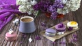 A row of colorful macaroon desserts on the table and a gray cup of coffee. Purple bouquet of lilac flowers. Royalty Free Stock Photo