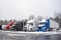 Row of the different big rigs semi trucks with loaded semi trailers standing on the winter truck stop parking lot with snow and Royalty Free Stock Photo