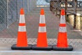 Row of three construction cones in urban area for construction or industry in late afternoon shade with chain link fence