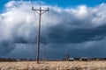 Row of wooden electricity poles with three wires and ceramic isolators in field. Heavy dark blue clouds before thunderstorm, brown Royalty Free Stock Photo