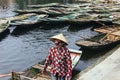 Rowing boat woman wearing red and white colors shirt, conical hat and mouth mask standing with empty boats over the river.