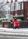 Royal Mail British post man in deep cold snow falling on road and icy frozen pavement delivering letters with trolley red uniform