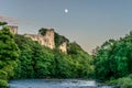 A ruin of a castle protruding through a forest next to a river with a moon