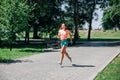 running smiling young sporty woman in park in sportswear on background of green trees on grey tiles in sunny summer day