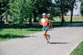running smiling young sporty woman in park in sportswear in front of green trees on road of grey tiles in sunny summer