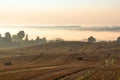 Rural colorful foggy landscape at sunrise. Harvested agricultural wheat field with straw bales and foggy countryside dale behind Royalty Free Stock Photo