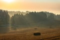 Rural colorful foggy landscape at sunrise. Harvested agricultural wheat field with straw bales and foggy forest behind it Royalty Free Stock Photo