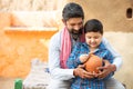 Rural Indian father and son putting coin into clay money box. Happy beard man and boy holding traditional piggy bank or Gullak,