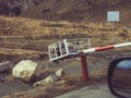 In a rural mountainous area among dry grass and boulders, a metal barrier gate weighted with stones blocks the passage