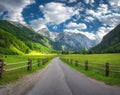 Rural road in alpine mountains, wooden fence, green meadows, sky