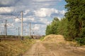 Rural sandy road between railroad with electric bearing pollars and pine forest belt with cloudy autumn sky on the background