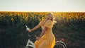 Rural woman in timeless dress riding retro styled white bicycle on country road alone near sunflowers field. Vintage Royalty Free Stock Photo