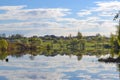 Russian village and green fields with trees and a blue sky with clouds reflected in the calm water of the lake. Calm and silence.