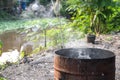 Rusted trash bin with burning garbage, smoke rising, set against a blurred riverside with wild grass Royalty Free Stock Photo