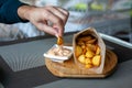 Rustic Fried potato wedges in paper envelope with sauce on wooden plate on the outdoor terrace in cafe. Mans hand takes slice and Royalty Free Stock Photo