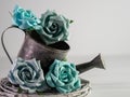 Rustic metal watering can with a gray wooden wreath and 4 teal and blue paper rose flower blooms and a soft gray background. Royalty Free Stock Photo