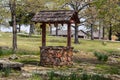 Rustic rock and wooden wishing well surrounded by paving stones and trees in springtime with log cabin in distance Royalty Free Stock Photo