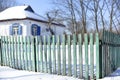 Rustic winter landscape with a wooden green fence and a snow-covered house behind it. A sunny day with a blue clear sky. Royalty Free Stock Photo