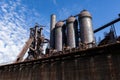 Rusting abandoned steel mill, blast furnaces and smokestacks atop a concrete wall, set against a brilliant blue sky